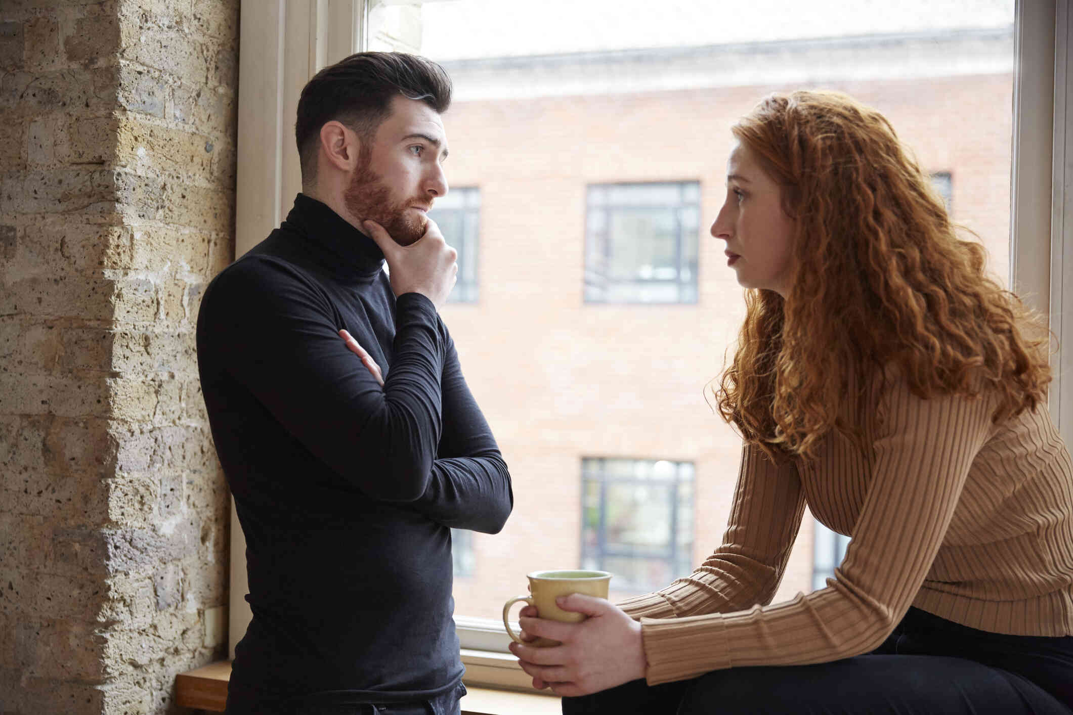 A man holds his chin, while a woman sits beside the window, holding her cup while looking eye-to-eye to her partner, the couple both look concerned.